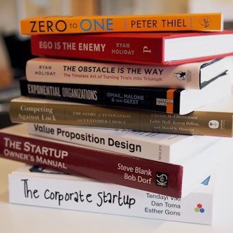 A stack of business and entrepreneurship books on a table. The titles are oriented towards strategies, innovation, and startup guidance, including 'Zero to One', 'Ego is the Enemy', and 'The Startup Owner's Manual'. The setting appears to be in a modern office or study environment.