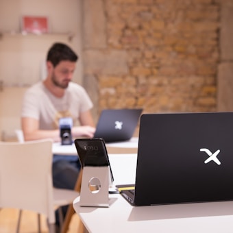 A modern workspace with a focus on technology. In the foreground, there is a black laptop with a distinctive logo placed on a table beside a smartphone propped on a stand. A man is sitting in the background, also using a similar laptop. The setting is minimalistic with a combination of white furniture and a textured brick wall.