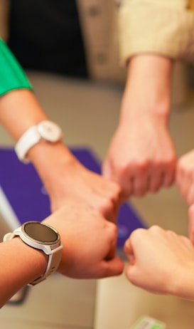 Several people form a circle, joining their fists together in a gesture of unity and teamwork. Various wristwatches and clothing styles are visible, suggesting a diverse group.